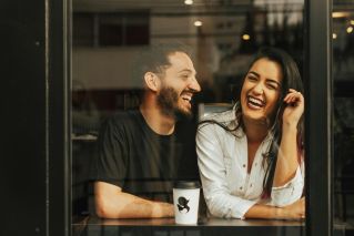A man and woman laughing together at a café window with coffee.
