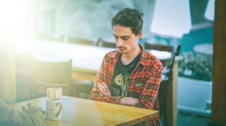 A young man sits at a cafe table, lost in thought, with a serene and contemplative expression.